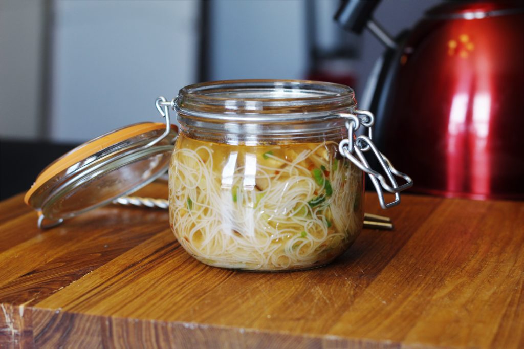 noodles and broth in a glass container. kettle in background.