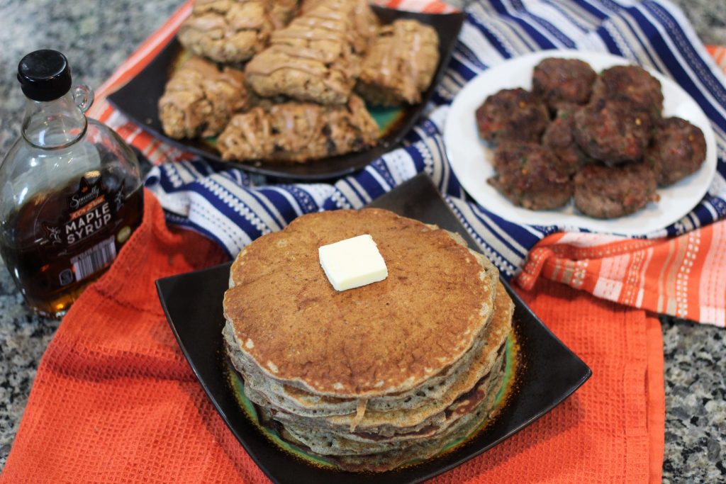 Pancakes stacked on a plate. Scones and sausages are in the background. 