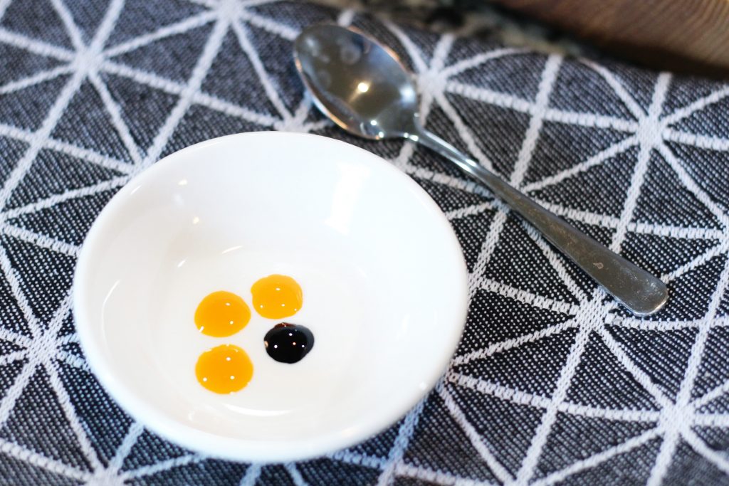 A little ceramic bowl with 3 drops yellow food coloring and 1 drop green.  A tiny spoon is in the background. 