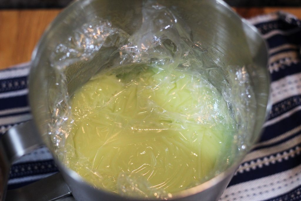 The ice cream base in a metal pitcher with plastic wrap pushed against the surface of the ice cream base. 