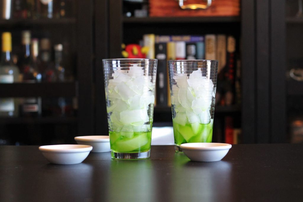 Two tall glasses filled with ice cubes, each with a small amount of melon liqueur at the bottom. 