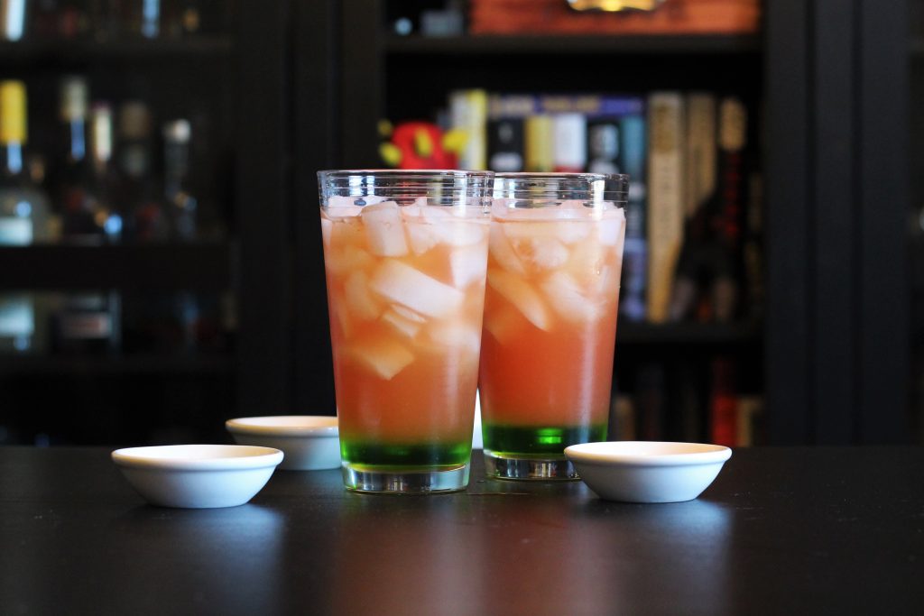 Two tall glasses filled with ice cubes, each with a small amount of melon liqueur at the bottom, and the watermelon mix on top.