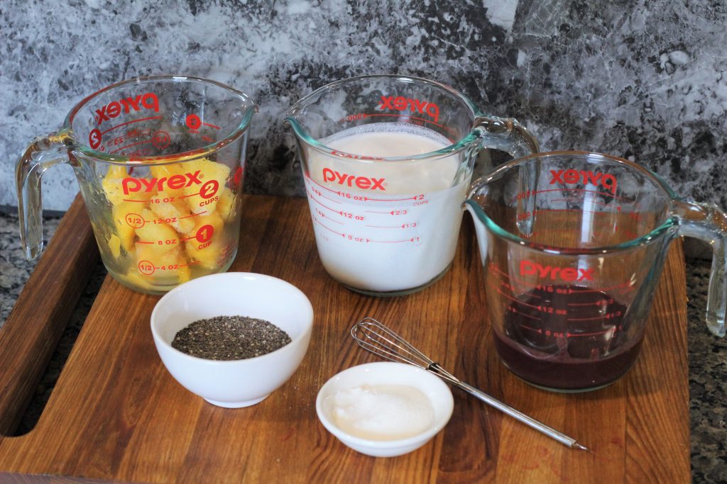 Clockwise from top left: measuring cup with mango chunks, measuring cup with coconutmilk, measuring cup with acai puree, small metal whisk, tiny bowl with sugar, small bowl with chia seeds