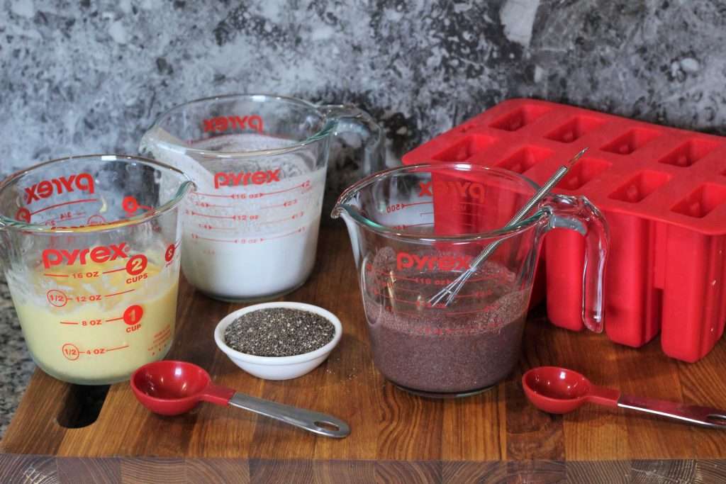 Left to right: measuring cup with blended mango coconut milk mix, measuring cup with coconut milk, tiny bowl with chia seeds, measuring cup with acai coconut milk mix, silicone popsicle mold