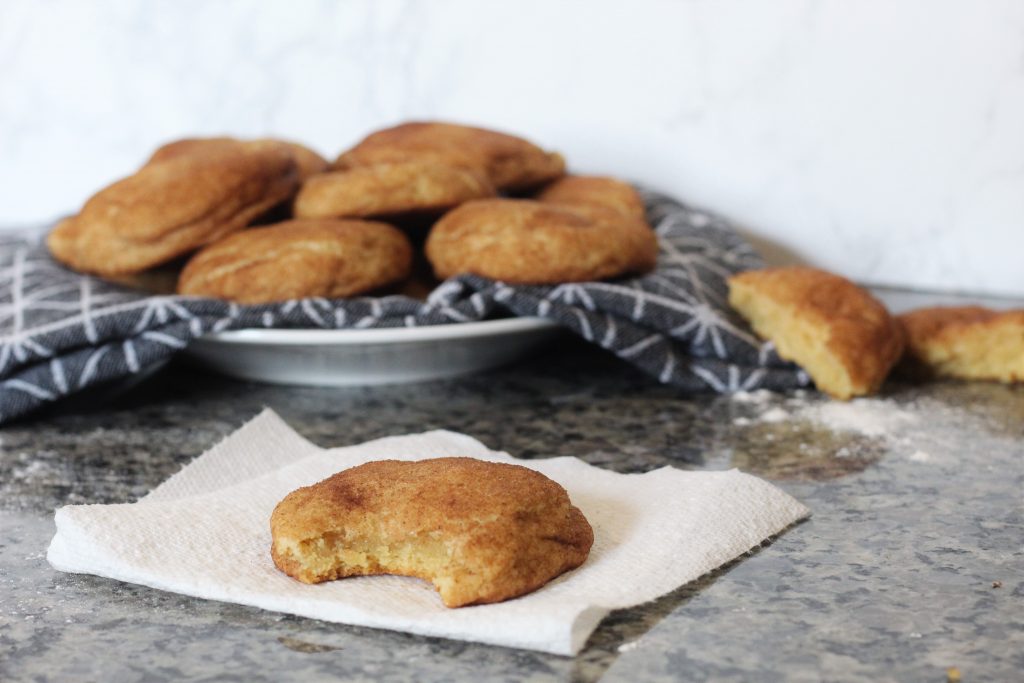 A cookie with a bite taken out on a paper towel. In the background is a plate with a cloth napkin with more cookies piled on it. 