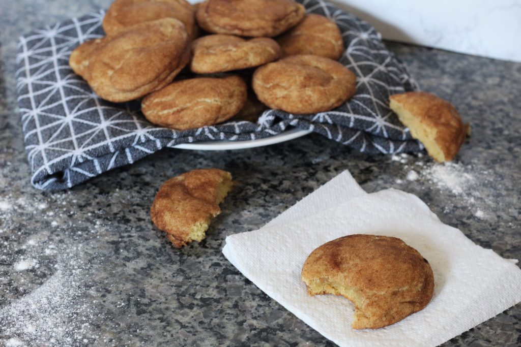 A cookie with a bite taken out on a paper towel. In the background is a plate with a cloth napkin with more cookies piled on it. 