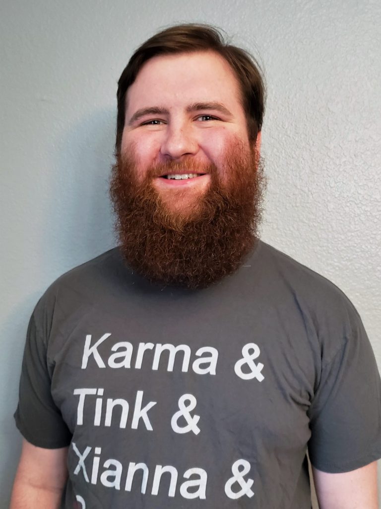 Photo of Hudson. He is a white man, and is smiling and wearing a Tabletop Squadron shirt that says "Karma & TInk & Xianna & Sabos" with a red line through Sabos. 
He has short brown hair, and a large auburn beard.