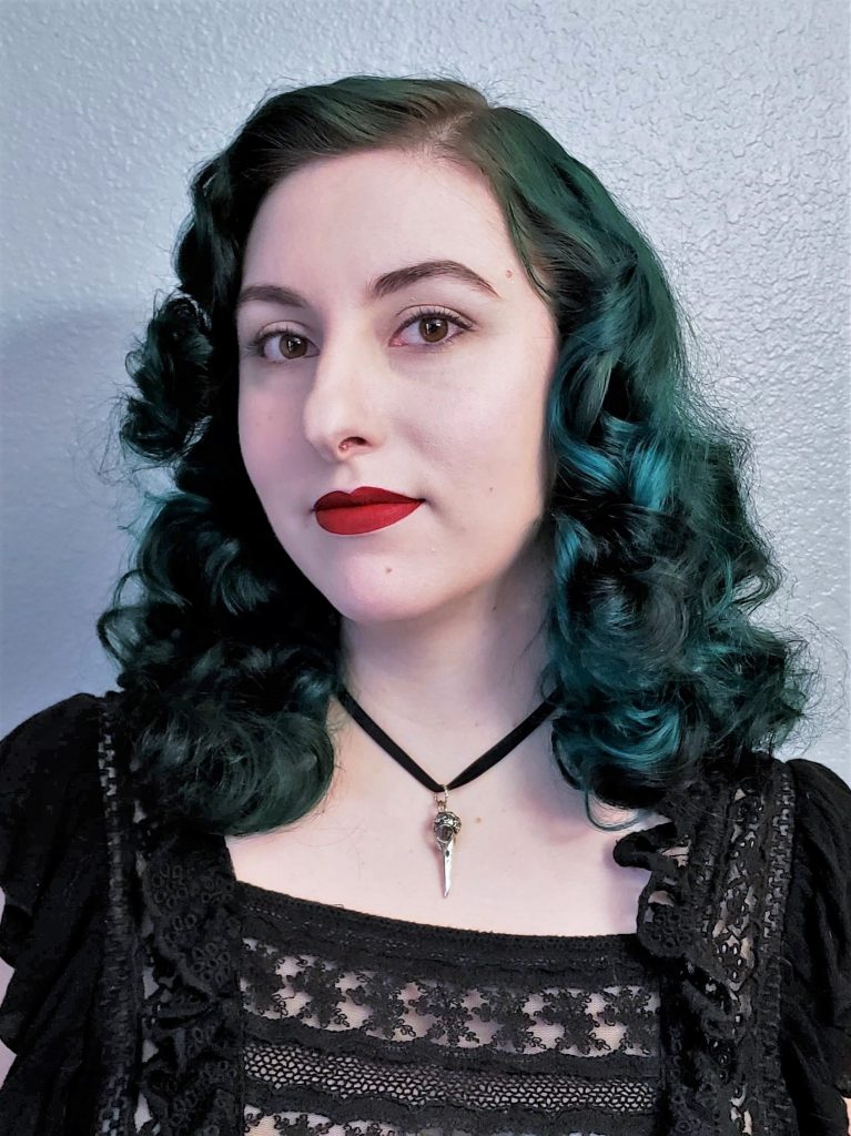 A photo of Laura. They are a white nonbinary person with dark teal hair that is curled in a vintage style. They are wearing a black top and small bird skull necklace. 