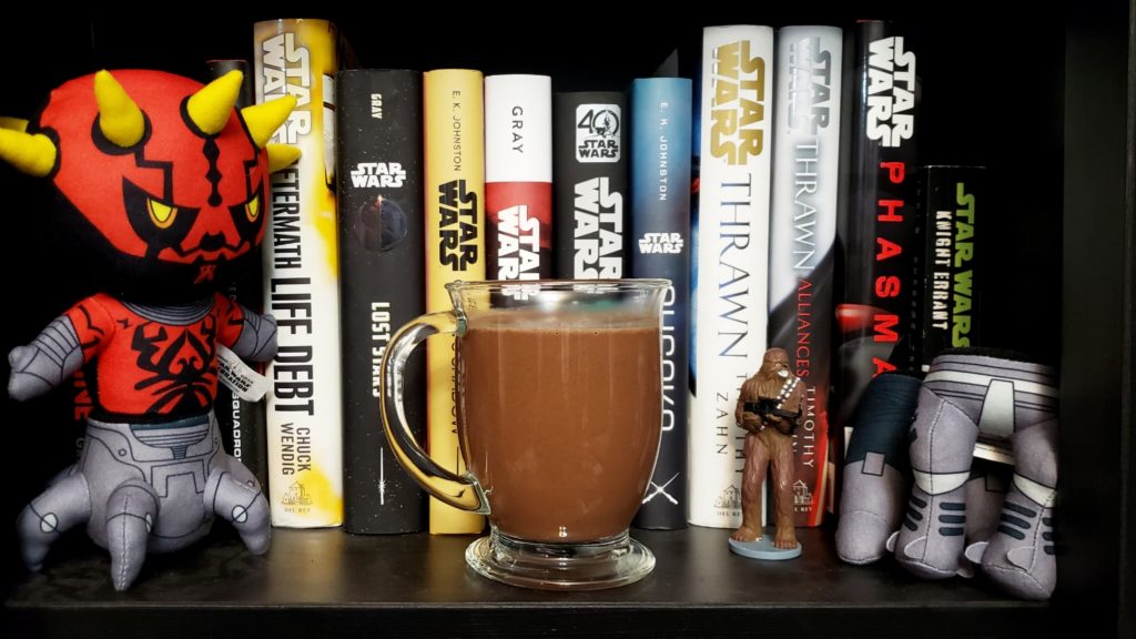 A clear mug of hot chocolate in front of Star Wars books on a shelf. To one side is a Darth Maul plushie, and on the other side is a small Chewbacca toy.